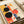 May include: Abstract geometric art print displayed in light wood frame on white surface next to cream-colored chair. The artwork features bold black circles, orange and red rectangular shapes, blue striped elements, and curved forms in a modernist composition. Warm natural lighting creates soft shadows on the neutral-toned setting.