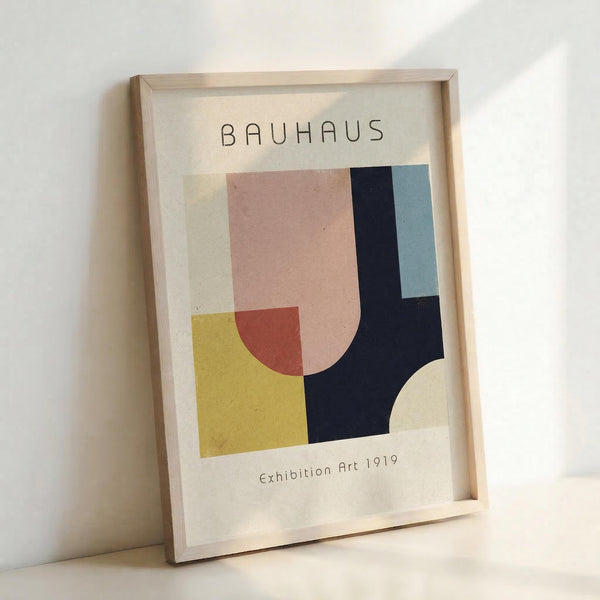 May include: Framed Bauhaus-inspired abstract art print leaning against white wall in light wood frame. Poster features geometric shapes in pink, yellow, red, navy blue, and light blue on cream background with "BAUHAUS" text at top and "Exhibition Art 1919" at bottom. Modern minimalist wall art mockup showing vintage-style design poster.