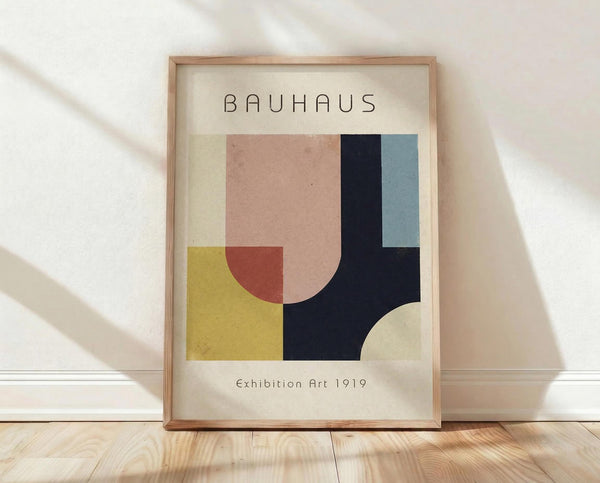 May include: Framed Bauhaus-style geometric abstract art print with overlapping shapes in muted tones of pink, yellow, navy blue, sage green, and cream. Modern minimalist poster displayed in natural wood frame, leaning against white wall on light wooden floor with soft natural lighting creating shadows.