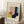 May include: Framed Bauhaus-style geometric art print leaning against white wall corner. Abstract composition features overlapping shapes in yellow, coral pink, navy blue, and light blue on cream background. Wooden frame sits on white textured rug beside wooden side table with white coffee cup. Natural window light creates soft shadows on wall.