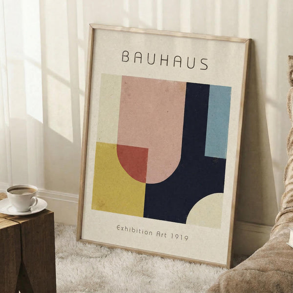 May include: Framed Bauhaus-style geometric art print leaning against white wall corner. Abstract composition features overlapping shapes in yellow, coral pink, navy blue, and light blue on cream background. Wooden frame sits on white textured rug beside wooden side table with white coffee cup. Natural window light creates soft shadows on wall.