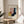 May include: Framed Bauhaus-style geometric art print with abstract shapes in yellow, coral pink, navy blue, and sage green colors, displayed on a light wood round side table next to a beige upholstered chair. Natural sunlight casts soft shadows across the minimalist room setting with white curtains and a plant visible in the corner.