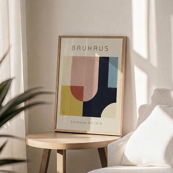 May include: Framed Bauhaus-style geometric art print with abstract shapes in yellow, coral pink, navy blue, and sage green colors, displayed on a light wood round side table next to a beige upholstered chair. Natural sunlight casts soft shadows across the minimalist room setting with white curtains and a plant visible in the corner.