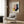 May include: Geometric abstract art print featuring overlapping shapes in pink, navy blue, yellow, and cream colors, displayed in light wooden frame on wood-paneled wall. Modern living room setting with beige upholstered armchair, geometric patterned throw pillow, brass floor lamp with marble base, and dark round side table with open book.