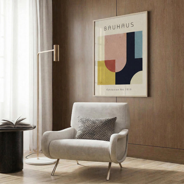 May include: Geometric abstract art print featuring overlapping shapes in pink, navy blue, yellow, and cream colors, displayed in light wooden frame on wood-paneled wall. Modern living room setting with beige upholstered armchair, geometric patterned throw pillow, brass floor lamp with marble base, and dark round side table with open book.