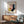 May include: Framed Bauhaus-style poster with geometric shapes in pink, yellow, navy, and teal displayed above mid-century modern walnut credenza with white ceramic lamp. Sage green upholstered armchair with wood frame sits nearby on hardwood floor. Natural sunlight casts shadows across white wall creating warm, minimalist living room scene.