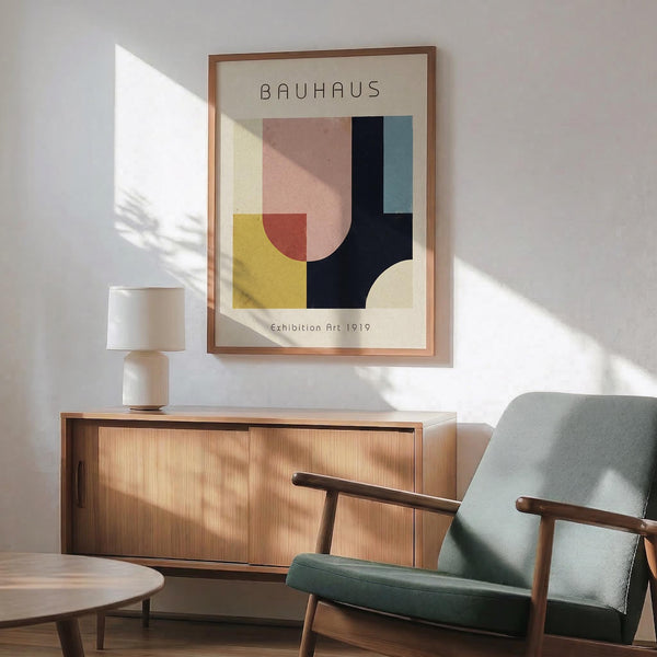 May include: Framed Bauhaus-style poster with geometric shapes in pink, yellow, navy, and teal displayed above mid-century modern walnut credenza with white ceramic lamp. Sage green upholstered armchair with wood frame sits nearby on hardwood floor. Natural sunlight casts shadows across white wall creating warm, minimalist living room scene.