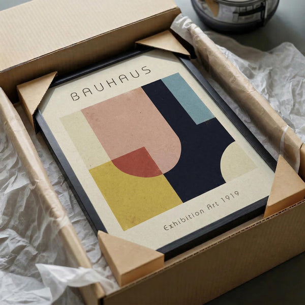 May include: Framed Bauhaus-style abstract art print with geometric shapes in pink, yellow, navy blue, and teal colors on cream background, displayed in black frame inside cardboard shipping box with protective tissue paper wrapping