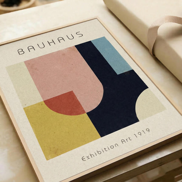 May include: Abstract geometric art print in wooden frame featuring overlapping shapes in pink, yellow, navy blue, and light blue with "BAUHAUS" and "Exhibition Art 1919" text. Frame sits against neutral backdrop with rolled paper tube beside it, showing vintage-inspired modernist design with clean lines and bold color blocks.