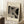 May include: A framed Bauhaus-style art print leaning against white paneled walls on a textured white rug. The poster features geometric black and striped shapes on cream paper with "BAUHAUS" and "Exhibition Art 1919" text. A wooden frame houses the vintage-inspired design. Nearby sits a white coffee cup on a saucer atop a rustic wooden block, with a beige knit throw draped in the corner, creating a cozy minimalist interior scene.