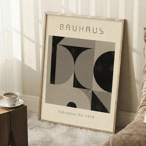 May include: A framed Bauhaus-style art print leaning against white paneled walls on a textured white rug. The poster features geometric black and striped shapes on cream paper with "BAUHAUS" and "Exhibition Art 1919" text. A wooden frame houses the vintage-inspired design. Nearby sits a white coffee cup on a saucer atop a rustic wooden block, with a beige knit throw draped in the corner, creating a cozy minimalist interior scene.
