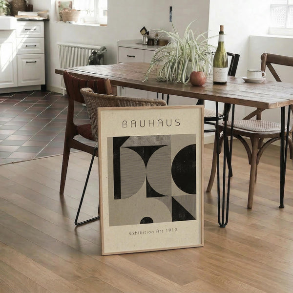 May include: Framed Bauhaus poster with geometric black shapes on cream background, displayed in modern kitchen dining area. Wood table with hairpin legs, wicker and wood chairs, spider plant, wine bottle, and white cup. Light wood floors, white cabinets visible in background.