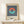 May include: Framed Bauhaus-style poster featuring coral and teal circular ring on navy and white vertical stripes, with "BAUHAUS Exhibition Art 1923" text below, displayed on white wooden dresser beside small wrapped gift with evergreen sprig against cream wall