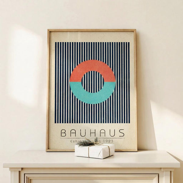 May include: Framed Bauhaus-style poster featuring coral and teal circular ring on navy and white vertical stripes, with "BAUHAUS Exhibition Art 1923" text below, displayed on white wooden dresser beside small wrapped gift with evergreen sprig against cream wall