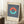 May include: Framed vintage-style Bauhaus poster with navy blue vertical stripes background, coral and turquoise split circle design, and beige text area. Light wood frame leaning against white wall corner next to beige textured chair and small wooden side table with white coffee cup and saucer on white fluffy rug.