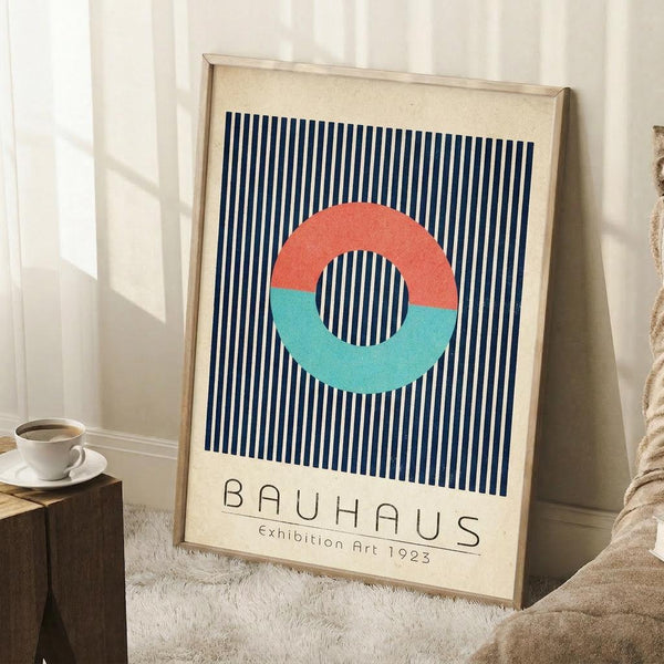 May include: Framed vintage-style Bauhaus poster with navy blue vertical stripes background, coral and turquoise split circle design, and beige text area. Light wood frame leaning against white wall corner next to beige textured chair and small wooden side table with white coffee cup and saucer on white fluffy rug.