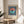 May include: Framed Bauhaus-style poster with coral and teal geometric circle on navy blue striped background, displayed on light wood round side table next to beige upholstered chair. Bright natural lighting creates shadows on white wall behind artwork. Green plant leaves visible in corner of minimalist modern interior setting.