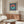 May include: Framed Bauhaus-style poster with coral and teal circle on navy striped background displayed above wooden sideboard in modern living room. White curved chair and glass oval coffee table visible. Poster features clean geometric design with exhibition text below colorful circle motif.