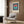 May include: Modern living room setting with beige armchair featuring geometric patterned pillow, gold floor lamp, dark side table with open book, and framed Bauhaus poster on wood-paneled wall. The poster displays a circular design in coral and teal colors over navy vertical stripes on cream background. Natural lighting from white curtained window illuminates the contemporary space with light wood flooring.