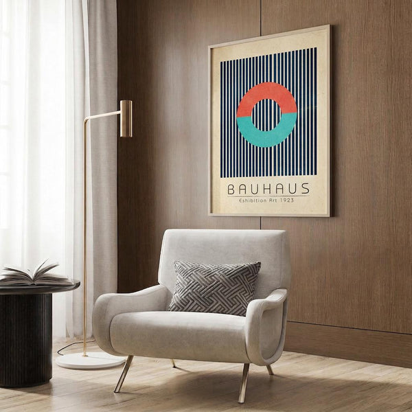 May include: Modern living room setting with beige armchair featuring geometric patterned pillow, gold floor lamp, dark side table with open book, and framed Bauhaus poster on wood-paneled wall. The poster displays a circular design in coral and teal colors over navy vertical stripes on cream background. Natural lighting from white curtained window illuminates the contemporary space with light wood flooring.
