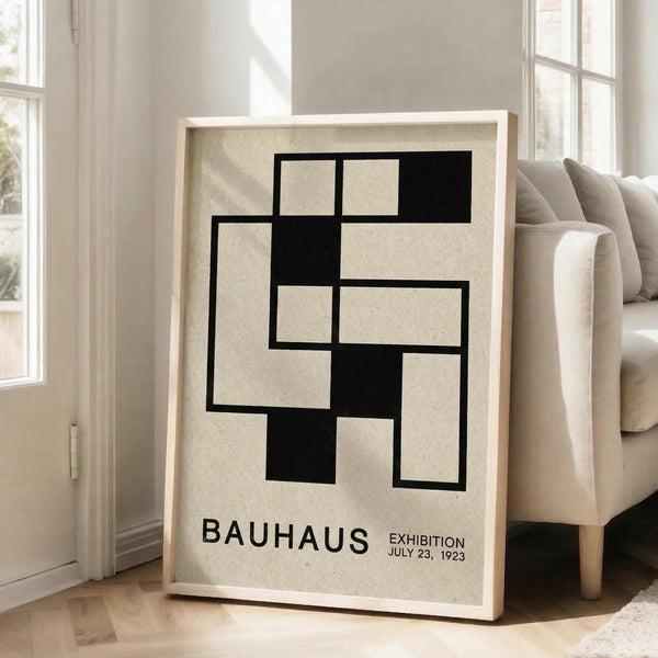 May include: Framed Bauhaus poster with geometric black rectangular shapes arranged in an abstract pattern on beige background, displayed in light wood frame leaning against white wall next to beige sofa in bright modern living room with large windows and natural lighting.