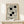 May include: Framed vintage-style Bauhaus exhibition poster with geometric black and white interlocking rectangles design, displayed on light wood shelf against cream wall. Small wrapped gift box with twine and greenery sprig sits beside the thin wood frame.