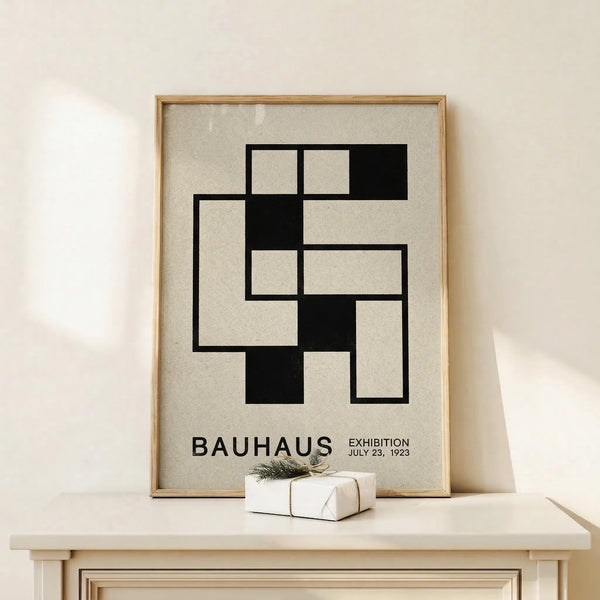 May include: Framed vintage-style Bauhaus exhibition poster with geometric black and white interlocking rectangles design, displayed on light wood shelf against cream wall. Small wrapped gift box with twine and greenery sprig sits beside the thin wood frame.