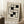 May include: A framed Bauhaus-style poster leaning against a white wall corner, featuring geometric black squares and rectangles arranged in an interlocking pattern on cream background. The minimalist artwork is displayed in a light wood frame, positioned next to a wooden block with white coffee cup and saucer, on a textured white rug beside a beige knitted throw blanket.