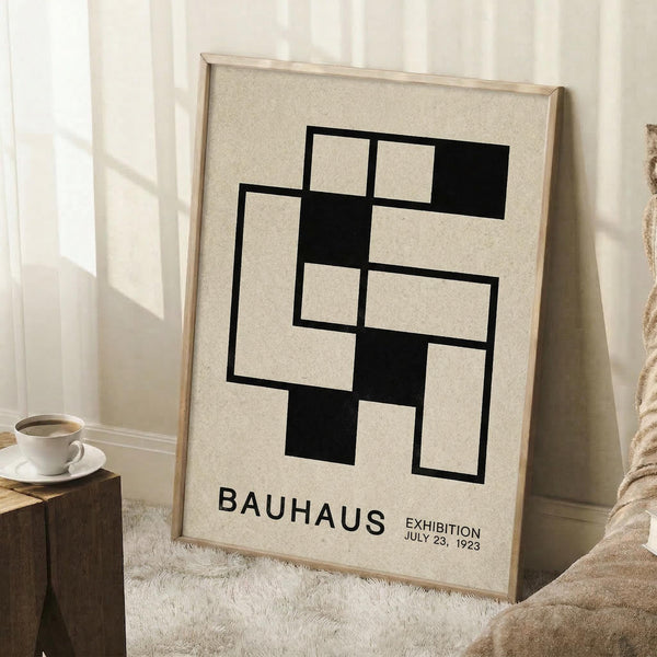 May include: A framed Bauhaus-style poster leaning against a white wall corner, featuring geometric black squares and rectangles arranged in an interlocking pattern on cream background. The minimalist artwork is displayed in a light wood frame, positioned next to a wooden block with white coffee cup and saucer, on a textured white rug beside a beige knitted throw blanket.
