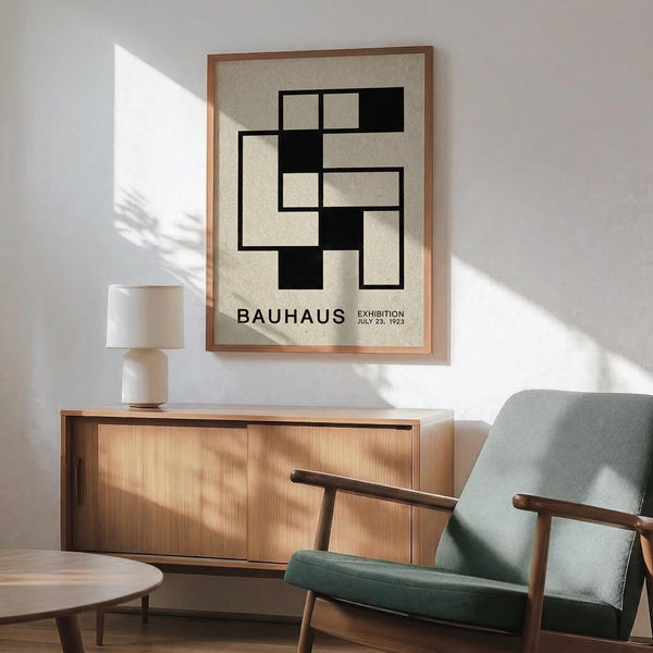 May include: Modernist black and white geometric poster with interlocking rectangular shapes and "BAUHAUS EXHIBITION JULY 23, 1923" text, displayed in thin copper frame above mid-century wooden credenza with white ceramic table lamp. Sage green upholstered armchair with wooden frame sits beside small round side table. Bright natural light casts shadows across white wall in minimalist room setting.