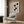 May include: Abstract geometric Bauhaus exhibition poster with black rectangular shapes on cream background, displayed in light wooden frame on warm wood-paneled wall. Modern cream upholstered armchair with geometric patterned pillow sits below, accompanied by brass floor lamp and dark round side table with open book. Soft natural lighting from window with beige curtains creates minimalist, mid-century modern room setting.