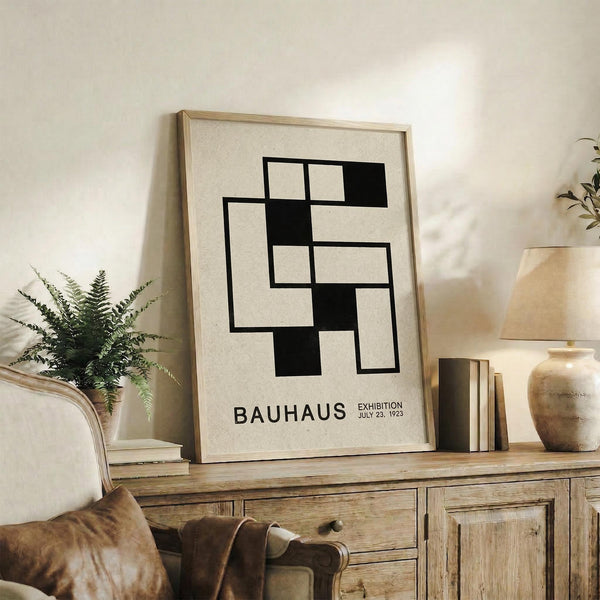 May include: Black and white geometric Bauhaus exhibition poster in light wood frame, displayed on rustic wooden sideboard with books, ceramic vase, and fern plant. Warm, neutral-toned living room setting with leather armchair visible. Poster features interlocking rectangular shapes in minimalist modernist style.
