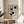 May include: A minimalist geometric poster with black rectangular shapes arranged in an abstract pattern on cream background, displayed in natural wood frame on round wooden side table next to beige upholstered chair. Soft natural lighting creates gentle shadows across neutral-toned room with white curtains and plant leaves visible in corner.
