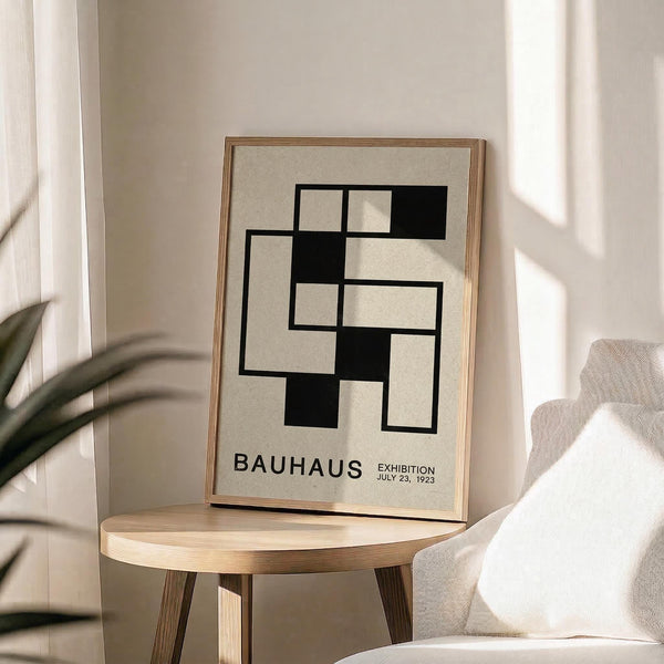 May include: A minimalist geometric poster with black rectangular shapes arranged in an abstract pattern on cream background, displayed in natural wood frame on round wooden side table next to beige upholstered chair. Soft natural lighting creates gentle shadows across neutral-toned room with white curtains and plant leaves visible in corner.