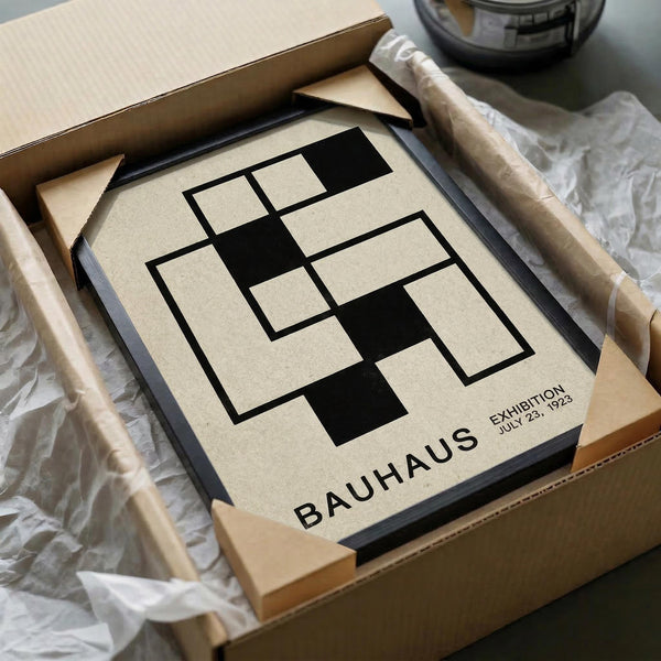 May include: Vintage Bauhaus exhibition poster with geometric black and beige design featuring interlocking rectangular shapes. The minimalist print sits in an open cardboard shipping box surrounded by white tissue paper and wooden dowels, creating an unboxing scene on a neutral surface.