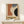 May include: Framed vintage-style Bauhaus poster with geometric shapes in rust orange, slate blue, cream, and black colors, displayed on light wood surface next to small white wrapped gift with twine and greenery sprig against neutral wall