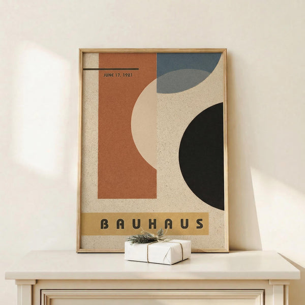 May include: Framed vintage-style Bauhaus poster with geometric shapes in rust orange, slate blue, cream, and black colors, displayed on light wood surface next to small white wrapped gift with twine and greenery sprig against neutral wall