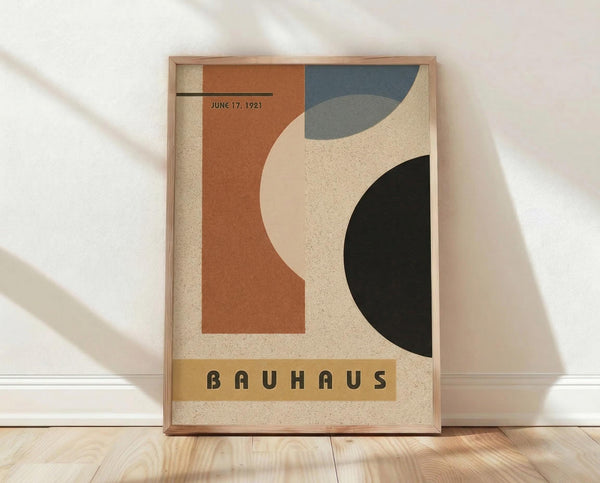 May include: Framed Bauhaus-style poster with geometric shapes in terracotta, navy blue, sage green, and black against cream background, displaying "JUNE 17, 1921" and "BAUHAUS" text. Wood frame leans against white wall on light wooden floor with soft natural lighting and fabric shadows.