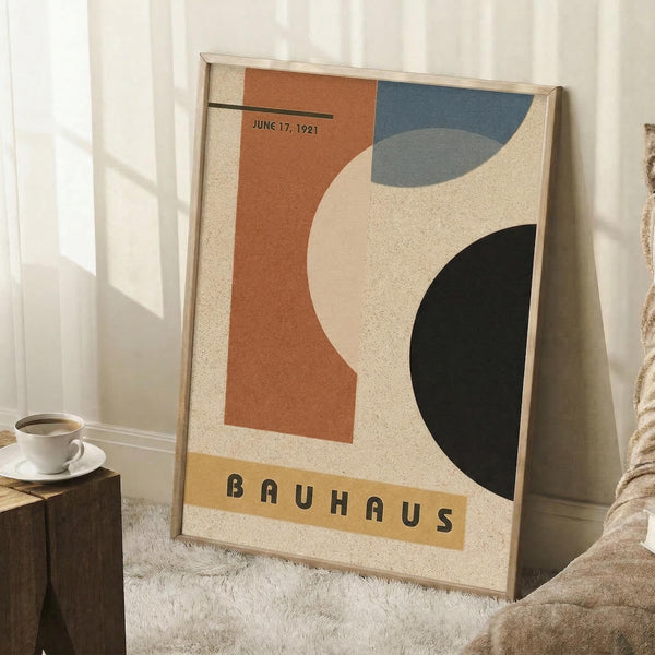May include: Framed abstract geometric poster with curved orange, blue, and black shapes on cream background, displaying "BAUHAUS" text and "JUNE 17, 1921" date. Wooden frame leans against white wall beside coffee cup on wooden block and textured throw blanket on white fuzzy rug.