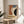 May include: Abstract modernist poster in wooden frame displaying geometric shapes in orange, blue, dark green and cream colors with "BAUHAUS" text and "JUNE 17, 1921" date. Frame sits on round wooden side table next to beige upholstered chair with plant leaves visible, soft natural lighting creating shadows on white wall background.