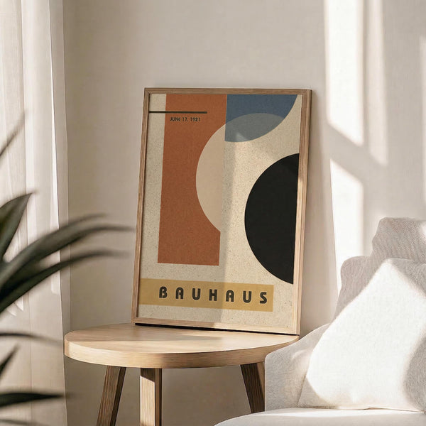 May include: Abstract modernist poster in wooden frame displaying geometric shapes in orange, blue, dark green and cream colors with "BAUHAUS" text and "JUNE 17, 1921" date. Frame sits on round wooden side table next to beige upholstered chair with plant leaves visible, soft natural lighting creating shadows on white wall background.