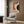 May include: Bauhaus-style poster with geometric shapes in orange, blue, cream and black colors, dated June 17, 1923, displayed in light wood frame on textured wood wall. Modern beige armchair with patterned pillow sits below, alongside brass floor lamp and dark round side table with open book. Neutral curtains visible on left.