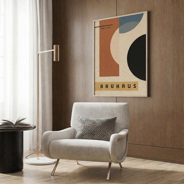 May include: Bauhaus-style poster with geometric shapes in orange, blue, cream and black colors, dated June 17, 1923, displayed in light wood frame on textured wood wall. Modern beige armchair with patterned pillow sits below, alongside brass floor lamp and dark round side table with open book. Neutral curtains visible on left.