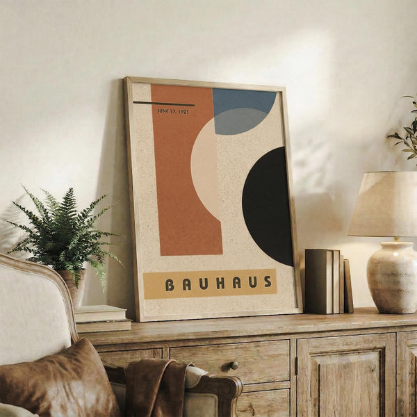 May include: Framed Bauhaus-style poster with abstract geometric shapes in terracotta orange, dusty blue, cream, and dark green, dated June 17, 1921. Displayed on rustic wooden credenza alongside fern plant, ceramic vases, and books in modern living room with neutral walls and leather furniture.