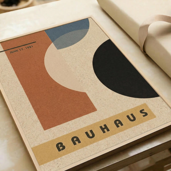 May include: Vintage Bauhaus exhibition poster from June 17, 1921 featuring abstract geometric shapes in terracotta orange, black, blue-gray, and cream against a textured beige background. The word "BAUHAUS" appears in black letters on a yellow banner at the bottom. The poster is displayed in an open book or portfolio alongside white paper materials on a neutral surface.