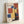 Framed vintage-style poster with geometric abstract composition in primary colors - red, blue, and yellow rectangular shapes arranged on cream background. Features "BAUHAUS" text at top and "1922" at bottom. Light wood frame leans against white wall, showing dimensional layered paper elements creating shadows and depth in the modernist design.