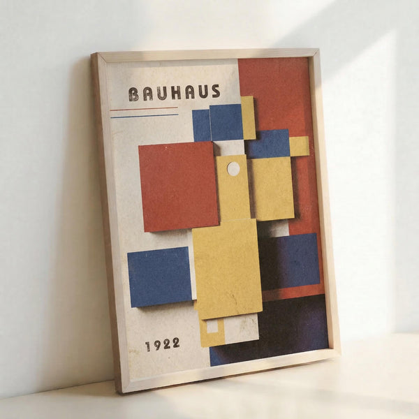 Framed vintage-style poster with geometric abstract composition in primary colors - red, blue, and yellow rectangular shapes arranged on cream background. Features "BAUHAUS" text at top and "1922" at bottom. Light wood frame leans against white wall, showing dimensional layered paper elements creating shadows and depth in the modernist design.