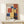 May include: Framed Bauhaus-style geometric art print with colorful overlapping rectangles in red, blue, yellow, and purple on cream background, displayed on light wood shelf next to small wrapped gift with evergreen sprig, against neutral wall with soft natural lighting