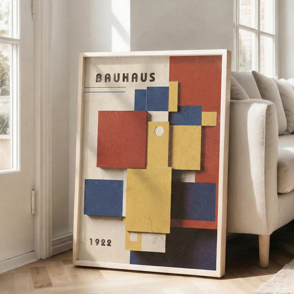 May include: Framed geometric art poster with "BAUHAUS 1922" text, featuring overlapping colorful squares and rectangles in red, blue, yellow, and white on cream background. Wooden frame leans against white wall next to beige upholstered chair in bright, minimalist living room with large windows and hardwood floors.