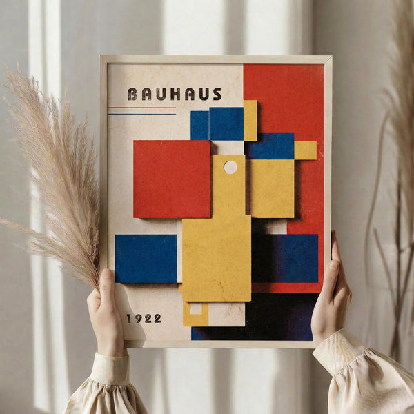 May include: Person holding framed Bauhaus poster with geometric composition of overlapping colored rectangles in red, blue, yellow, and white against cream background. Wood frame, neutral clothing, dried pampas grass visible, soft natural lighting near window with curtains.